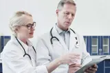 Twee artsen in witte jassen bespreken medische informatie op een tablet in een klinische omgeving.